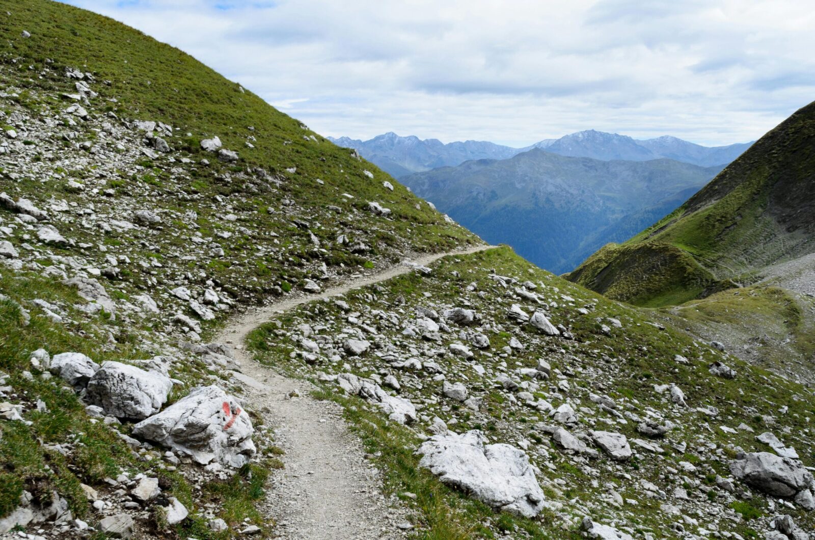 A winding trail through a rocky, lush green mountain landscape under a cloudy sky.